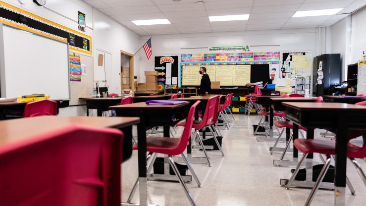 A teacher wearing a mask walks in an empty classroom with rows of desks and chairs.
