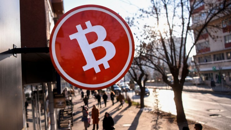 Pedestrians walk under a circular sign with a bitcoin logo.