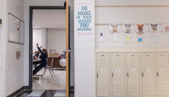 Through an open classroom door, two students raise their hands as they sit at their desks. The hall outside the classroom is visible, with lockers, student artwork and a sign that reads, "Do more of what makes you awesome."