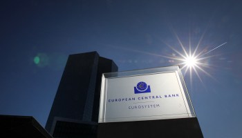 The headquarters of the European Central Bank is seen behind its sign. The sun is shining in the background amid a clear blue sky.