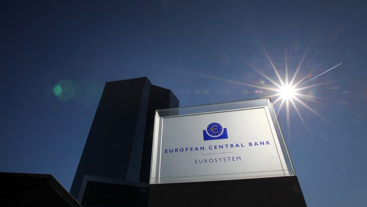 The headquarters of the European Central Bank is seen behind its sign. The sun is shining in the background amid a clear blue sky.