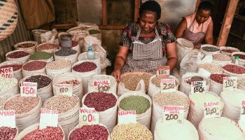 Two women wearing aprons sit behind rows of open bags of different colored cereals. The bags in front have prices and labels, reading baazi, rose-coco, royal, wairimu, yellow, pishori, kamande, basmati and long grain.