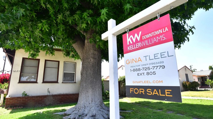 A single-story bungalow house with a large, shady tree in its front yard is seen behind a for sale sign.