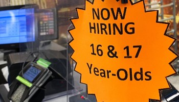 Signage advertising now hiring for 16 and 17 year old employees is displayed on a cash register inside a discount department retail store.