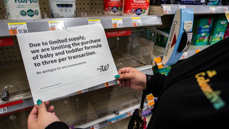 A woman holds a sign hanging from a pharmacy store shelf that explains there is a limit on the amount of baby formula someone can buy because of the nationwide shortage.