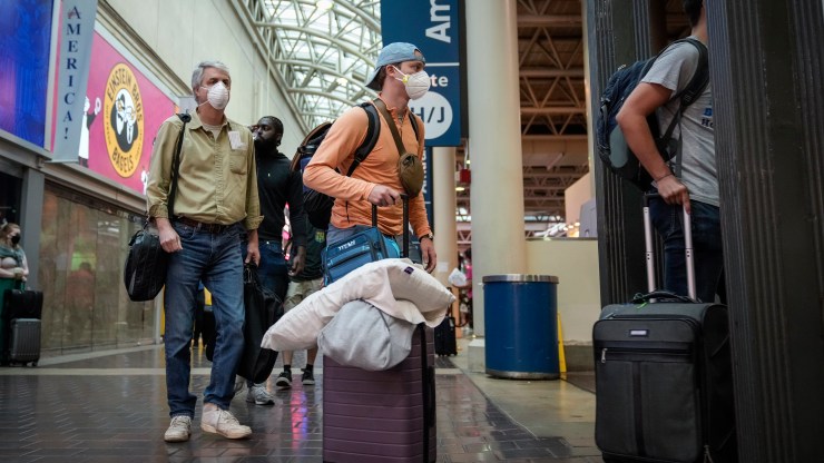 Masked travelers wait to board a train with luggage.