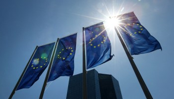 Flags of Europe fly in front of the European Central Bank as the sun shines behind.