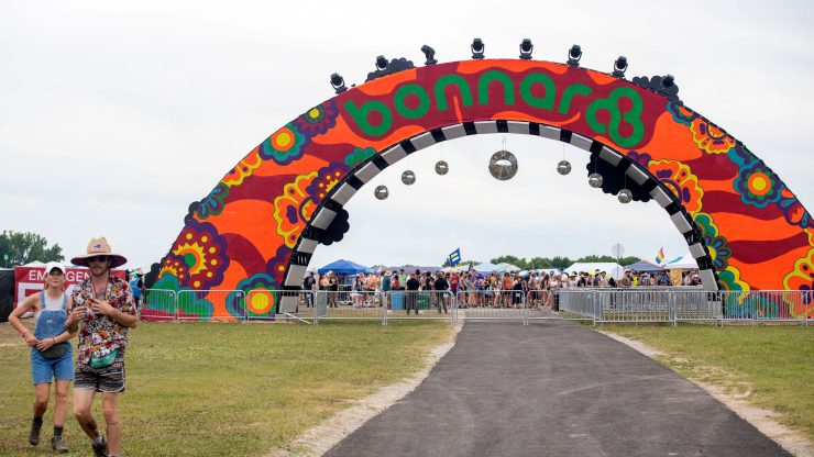 A large orange archway reads "Bonnaroo" in green text. Festival-goers stand beneath the arch, behind metal fences.