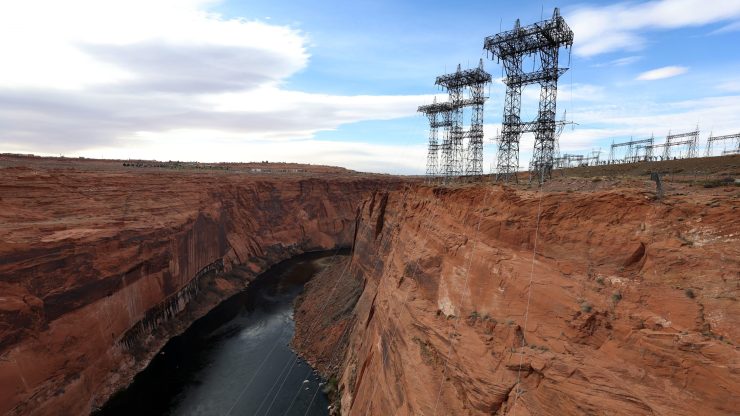 Several power line structures rise over a cliff face and the Colorado River. The river level is extremely low.