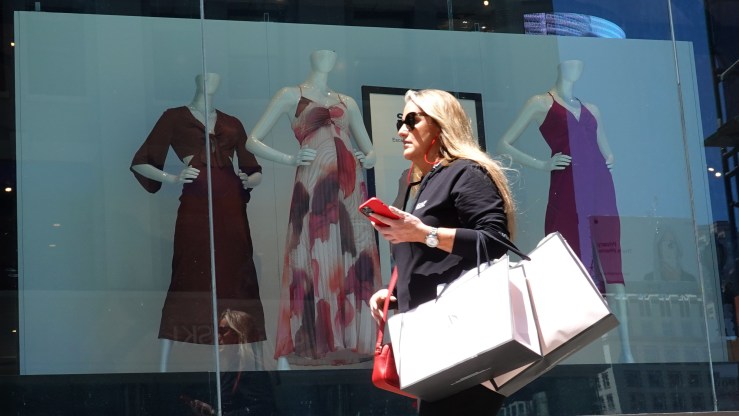 A woman with phone in hand and shopping bags on her arm passes mannequins in a shop window.