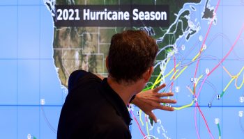NOAA National Hurricane Center Deputy Director Jamie Rhome stands with his back facing the camera in front of a map of the United States. Several yellow and pink lines on the East Coat show the path of a potential tropical storm, and Rhome uses his hand to show how the storm moves.