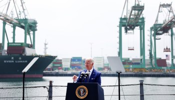 President Joe Biden delivers remarks in front of a presidential podium at the Port of Los Angeles. Cargo ships are seen in the background.