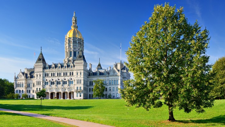 The Connecticut State Capitol in Hartford, Connecticut is seen in front of a blue sky and behind green grass and a full tree.