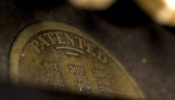 A closeup of the top half of a bronze-colored patent stamp. The visible text says "Patented."