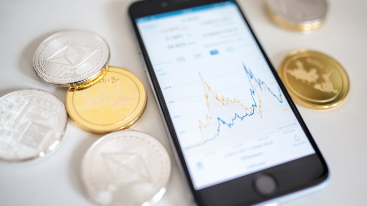 A phone rests next to litecoin, ripple and ethereum cryptocurrency altcoins. The smartphone displays a cryptocurrency price chart.