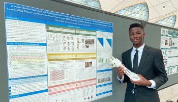 Okezue Bell, 16, stands next to an informational poster about the development of his prosthetic arm, which he is holding.