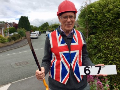 Tony Bennett wears a hard hat and union jack vest while holding a crowbar and road sign that reads '6'7"'.