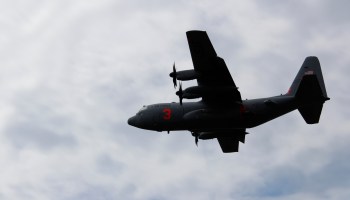 A military C-130 Hercules aircraft flies in a cloudy sky.