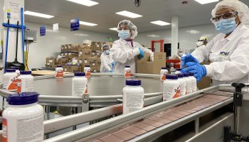 Workers in white jackets, hairnets, masks and gloves work at a pill processing conveyer belt.