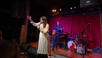 Artist Tristen Gaspadarek performs a farewell show at Mercy Lounge in Nashville. She stands at a microphone in a blouse and long skirt, in front of her band, which stands in front of red velvet curtains.