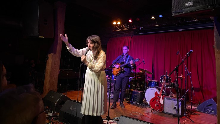 Artist Tristen Gaspadarek performs a farewell show at Mercy Lounge in Nashville. She stands at a microphone in a blouse and long skirt, in front of her band, which stands in front of red velvet curtains.