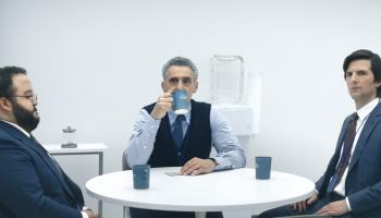 From left to right, actors Zach Cherry, John Turturro and Adam Scott in the show "Severance" sit around a table in the break room at an office with coffee mugs.