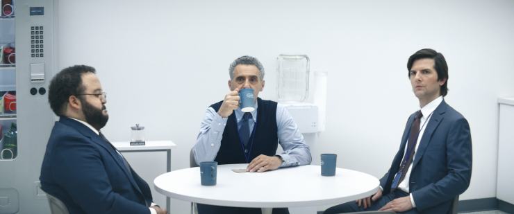 From left to right, actors Zach Cherry, John Turturro and Adam Scott in the show "Severance" sit around a table in the break room at an office with coffee mugs.