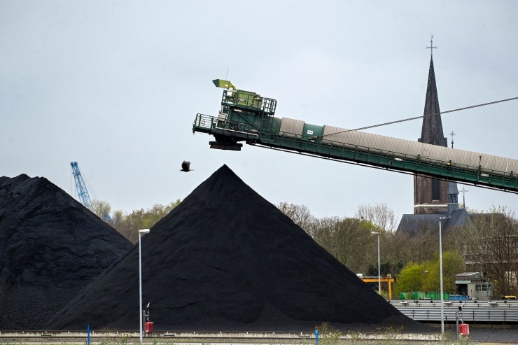 A crane hovers over pyramid-shaped piles of coal, with a church spire in the background.