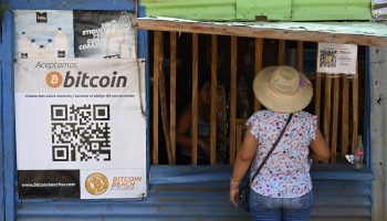 A woman with her back turned to the camera stands in front of a shopkeeper's storefront. She wears a wide-brimmed hat. In the left side of the image, there is a QR code and a sign that reads, "bitcoin."