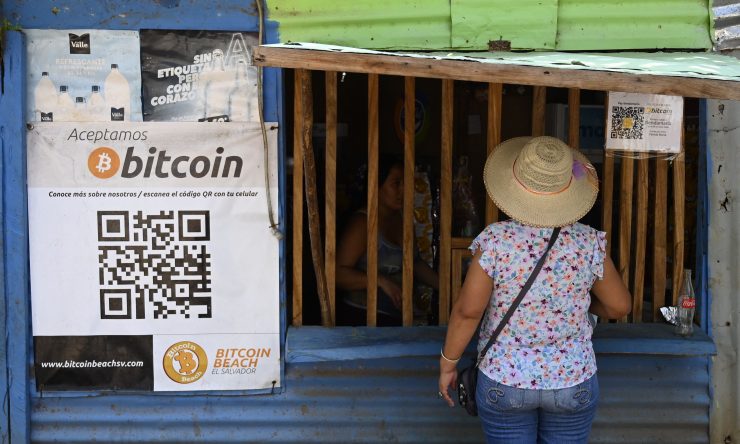 A woman with her back turned to the camera stands in front of a shopkeeper's storefront. She wears a wide-brimmed hat. In the left side of the image, there is a QR code and a sign that reads, "bitcoin."