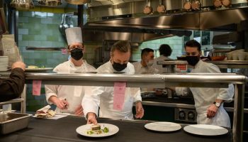 Chefs stand in a kitchen, facing the camera, plating food