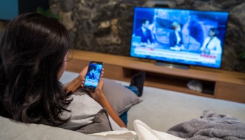 A woman sits on a couch, watching the news on TV and viewing her smartphone.