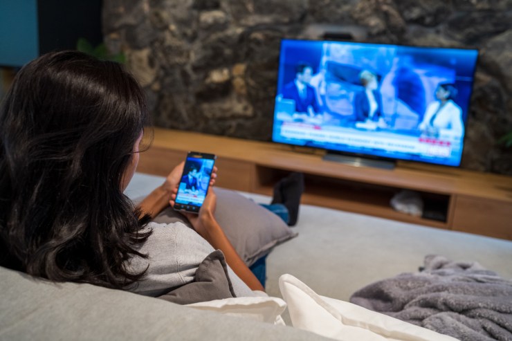 A woman sits on a couch, watching the news on TV and viewing her smartphone.