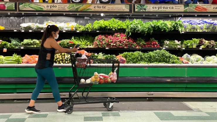 A customer pushes a cart through a fresh produce aisle at a grocery store.
