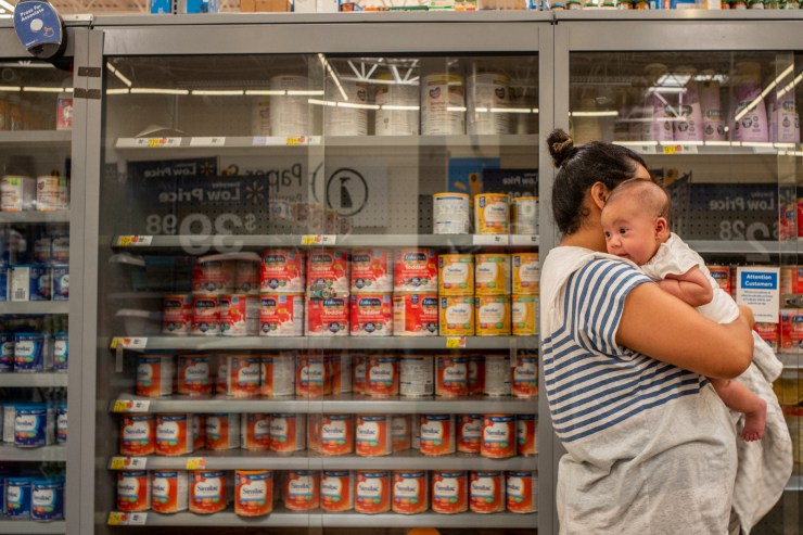 A woman holds a baby in a grocery store aisle. Behind them, a partially empty shelf for baby formula.