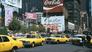 Taxis and pedestrians in front of massive advertisements looking toward Times Square in New York.