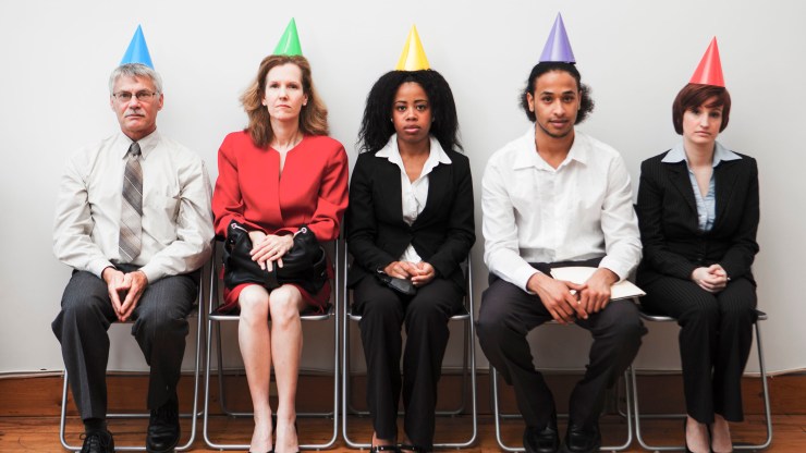 A group of business colleagues wearing party hats and semi-formal clothing, sitting awkwardly at an office party.