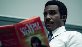 The character Milchick in the show "Severance," played by Tramell Tillman, holds up a self-help book and stares into the distance.