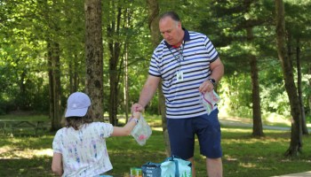 A man in shorts and a T-shirt hands a bag to a child wearing a baseball cap backward. They are surrounded by trees.