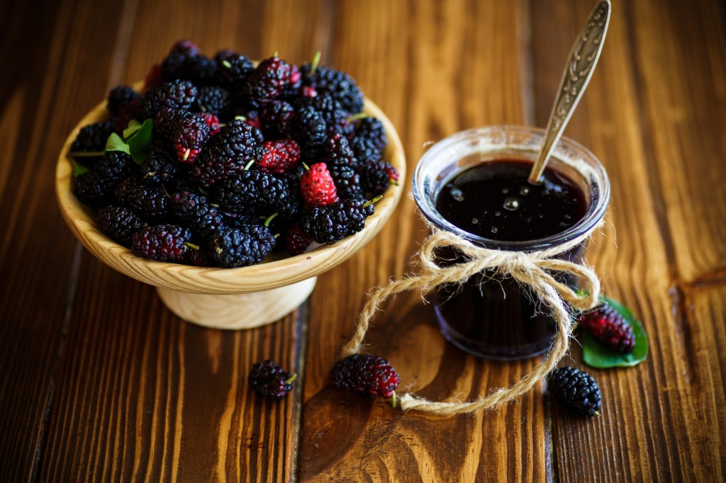 Mulberries, along with a glass cup of jam made out out of the fruit, sit on a wooden table.