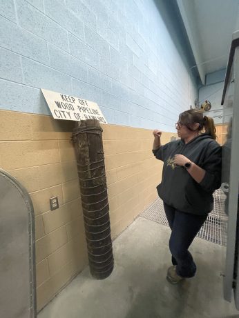 A woman stands in a narrow hallway across from a wooden pipe. A sign above the pipe says " Keep Off Wood Pipeline City of Rawling"