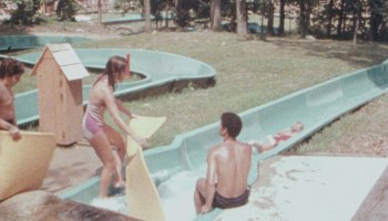 A girl gets ready to set down her mat at the top of a water slide, while a water park employee sits nearby.