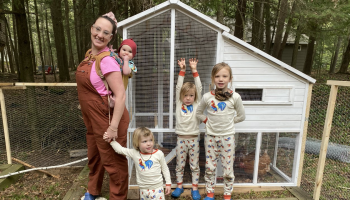 A mom stands with her four children in front of a chicken coop. One kid is strapped to the mom’s back, one is holding her hand and the other two are standing off to the right.