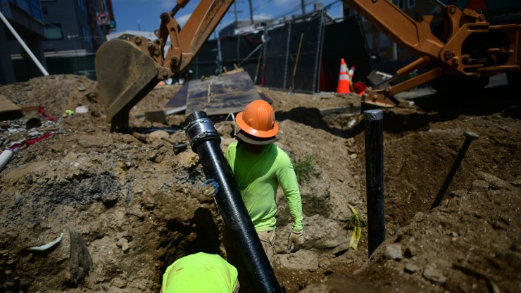 A construction worker stands in a ditch and holds a pipe while the sun beats down. There is a backhoe excavation machine in the background.