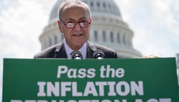 Senate Majority Leader Chuck Schumer stands in front a sign reading "Pass the Inflation Reduction Act." The Capitol dome is behind him.