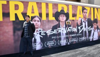 An actor stands in front of a Reservation Dogs banner at an event.