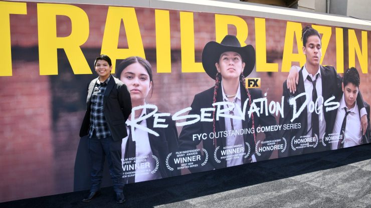 An actor stands in front of a Reservation Dogs banner at an event.