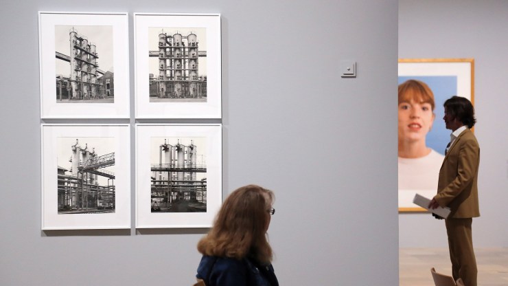 A woman sits in front of four black-and-white photographs of oil tanks taken by Bernd and Hilla Becher in a museum.