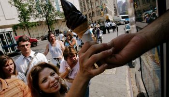 A woman reaches for an ice cream cone from a truck. It's a hot day and a line forms behind her.