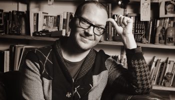 A black and white portrait shows Cory Doctorow smiling with his lips closed, his elbow bent and his thumb on his head. He sits in a leather chair in front of a crowded bookcase.